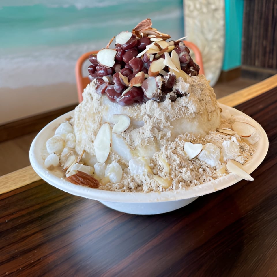 Hawaiian shave ice topped with red beans, almonds, and a light brown powder, on a wooden table.