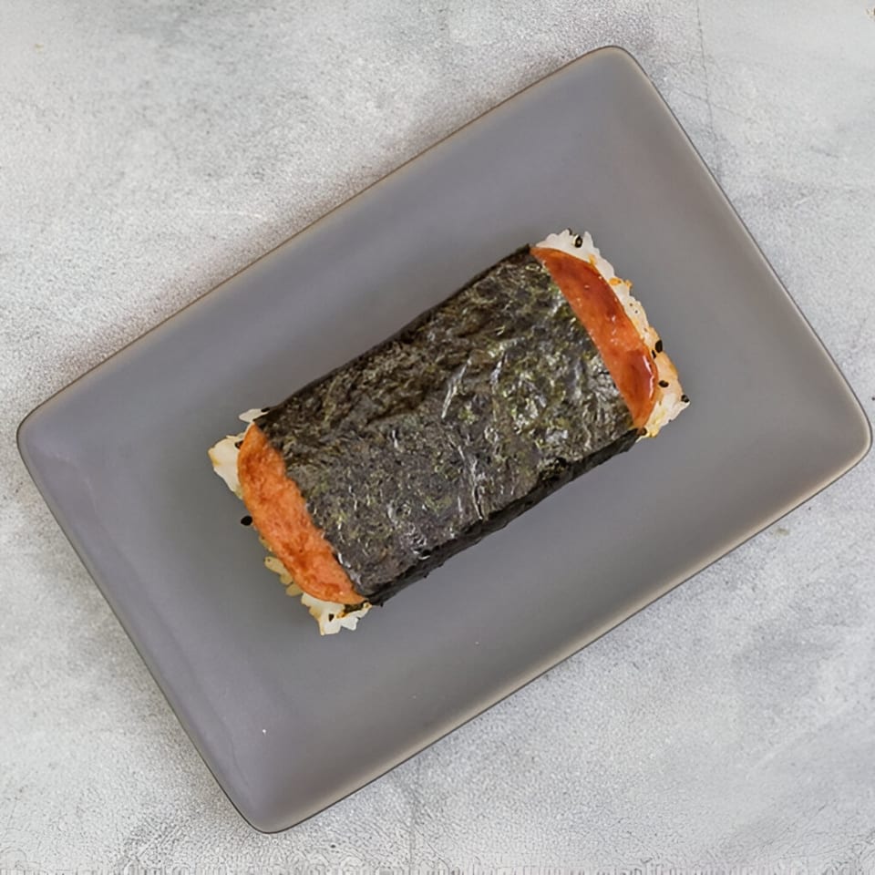 A Hawaiian Spam Musubi, a block of rice topped with grilled Spam and wrapped in nori, on a gray plate.