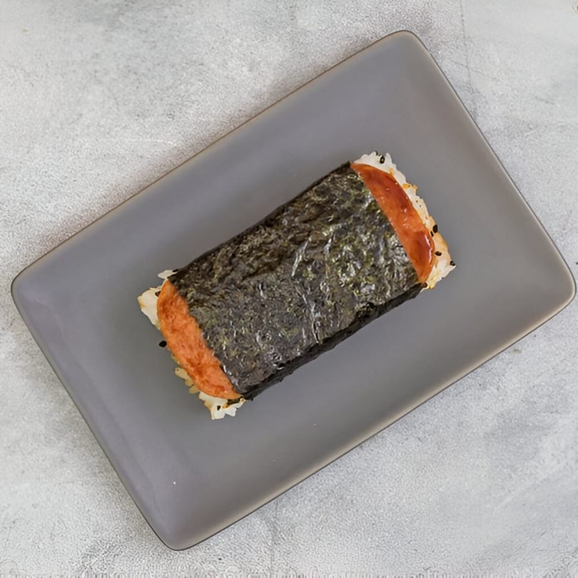 A Hawaiian Spam Musubi, a block of rice topped with grilled Spam and wrapped in nori, on a gray plate.